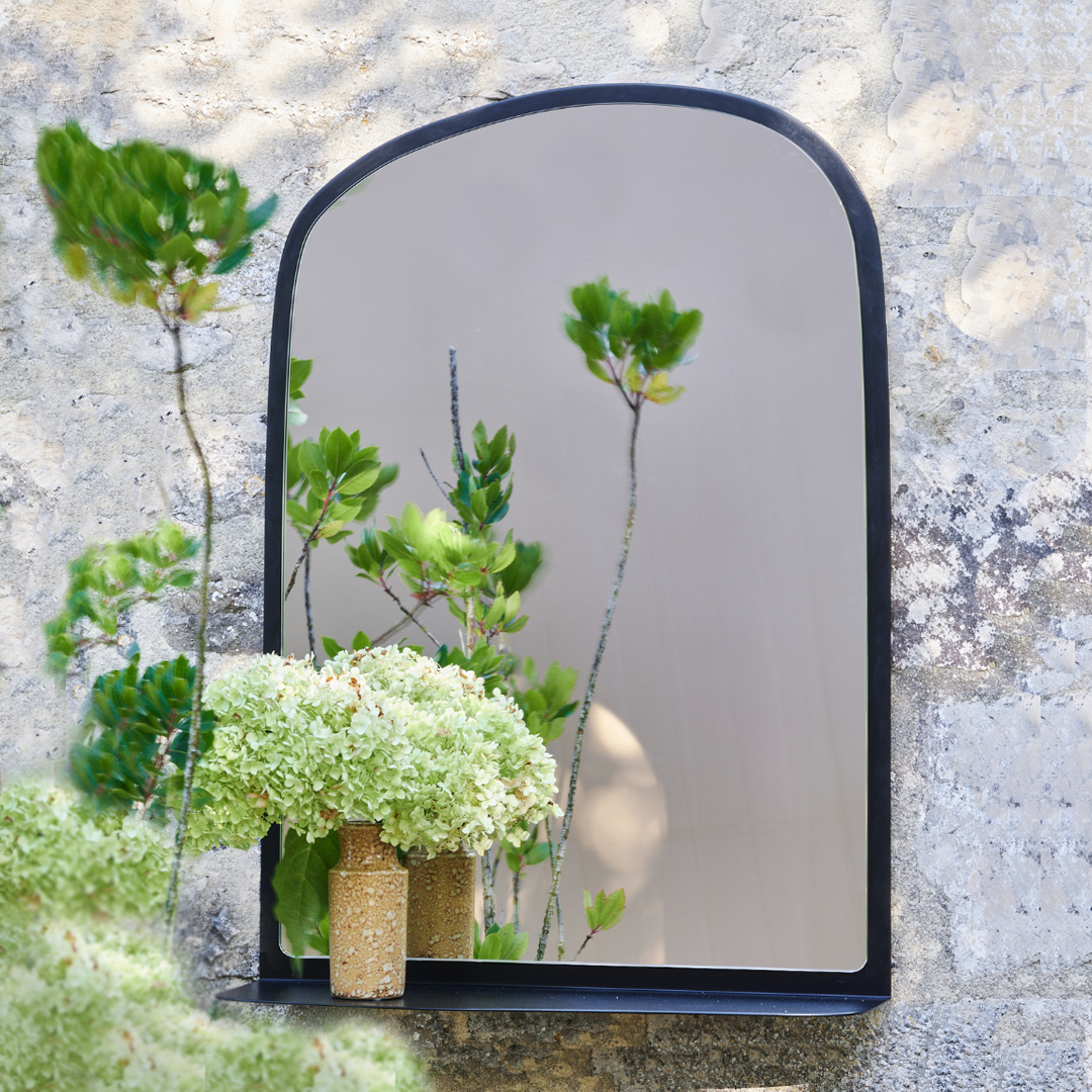 Miroir Aphrodite en acier, design épuré, entouré de fleurs dans un jardin moderne. Fabrication française artisanale.