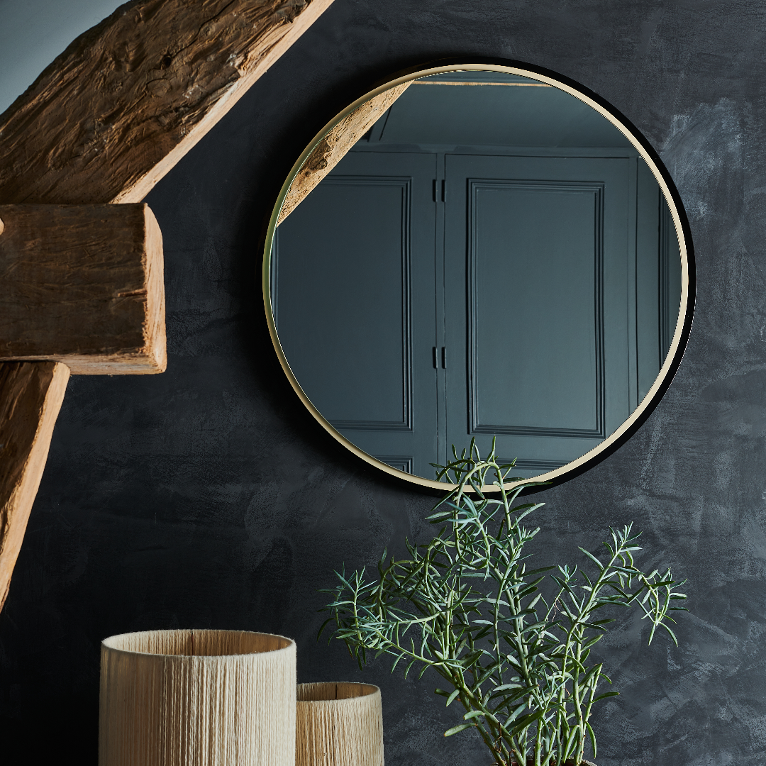 Miroir rond avec cadre doré, accroché sur un mur noir, dégageant une ambiance moderne et épurée. Plantes et vases en arrière-plan.