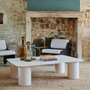 Table basse Métis en acier blanche, décorée de bouteilles et livres, dans un intérieur moderne avec mur en pierre.