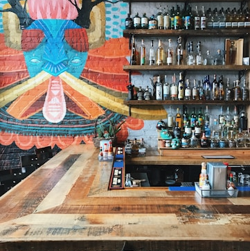 Bar avec un comptoir en bois, étagères garnies de bouteilles et mur coloré au design artistique. Atmosphère moderne et accueillante.