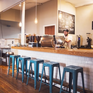 Bar d'un café moderne avec comptoir en bois, tabourets en métal bleu et barista préparant des boissons derrière la machine à café.