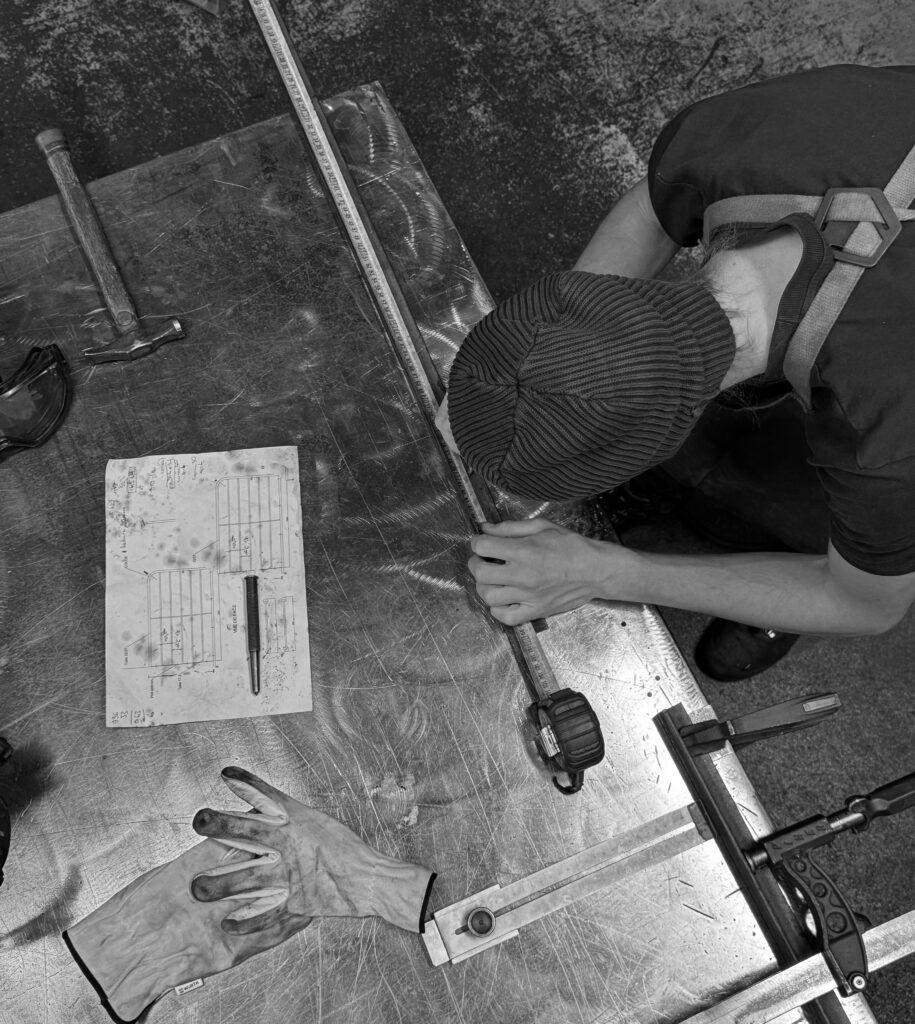 Artisan mesurant une pièce de métal dans l'atelier, avec des outils et des gants. Ambiance de création artisanale.